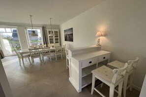 A modern kitchen with white cabinetry, a dining area with white chairs, and a view of the outdoors through large windows. - Holiday home with 4 bedrooms (Tetenbüll)