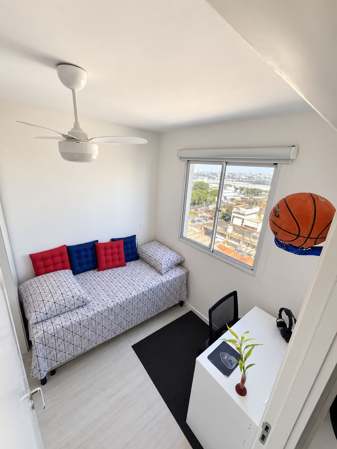 Quarto compacto com cama, escrivaninha, ventoinha e janela com vista para a cidade.