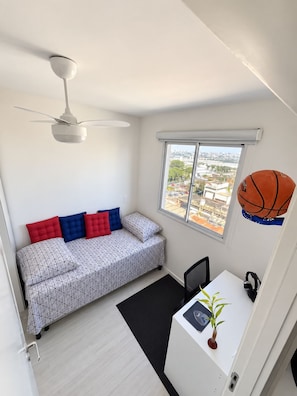 Una habitación pequeña con cama, escritorio, ventilador de techo y ventana con vista a la ciudad.