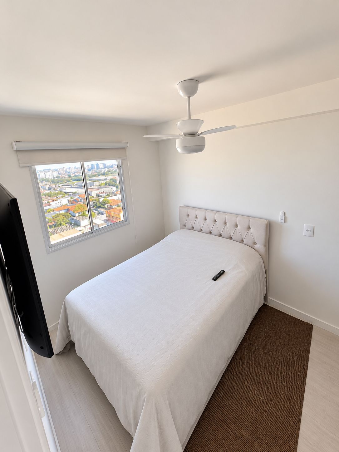 Quarto com cama, ventoinha no teto, janela com persianas e vista para edifícios.
