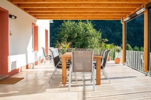 Eine Terrasse im Freien mit einem Holztisch und -stühlen, umgeben von Kübelpflanzen und Blick auf ein bewaldetes Gebiet.