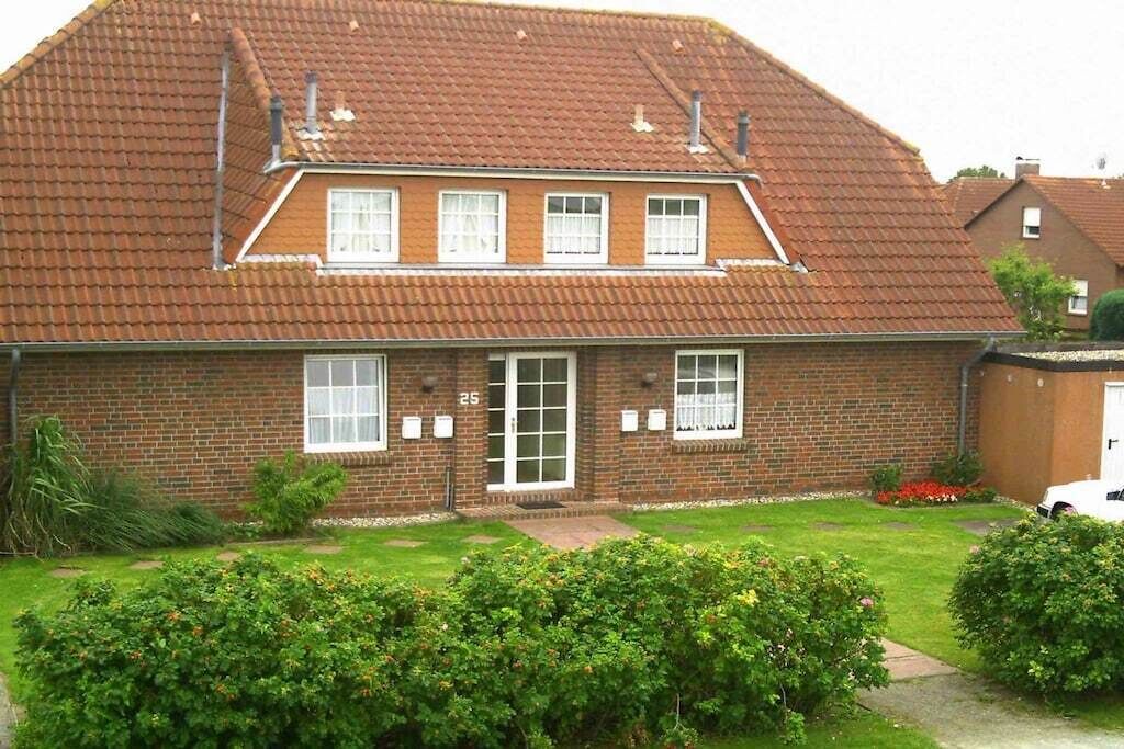 Une maison à deux étages, construite en briques, avec un toit en tuiles, une porte d’entrée et un petit jardin agrémenté d’arbustes et de fleurs.