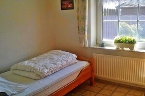A small, single-bed room with a wooden headboard, a radiator, a window with a view of a fence and greenery, and a potted plant on the windowsill. - Holiday apartment with 2 bedrooms (Neßmersiel)