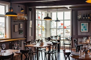 Ein Restaurantinneres mit mehreren Esstischen, Stühlen, hängenden Pendelleuchten und Blick durch große Fenster.
