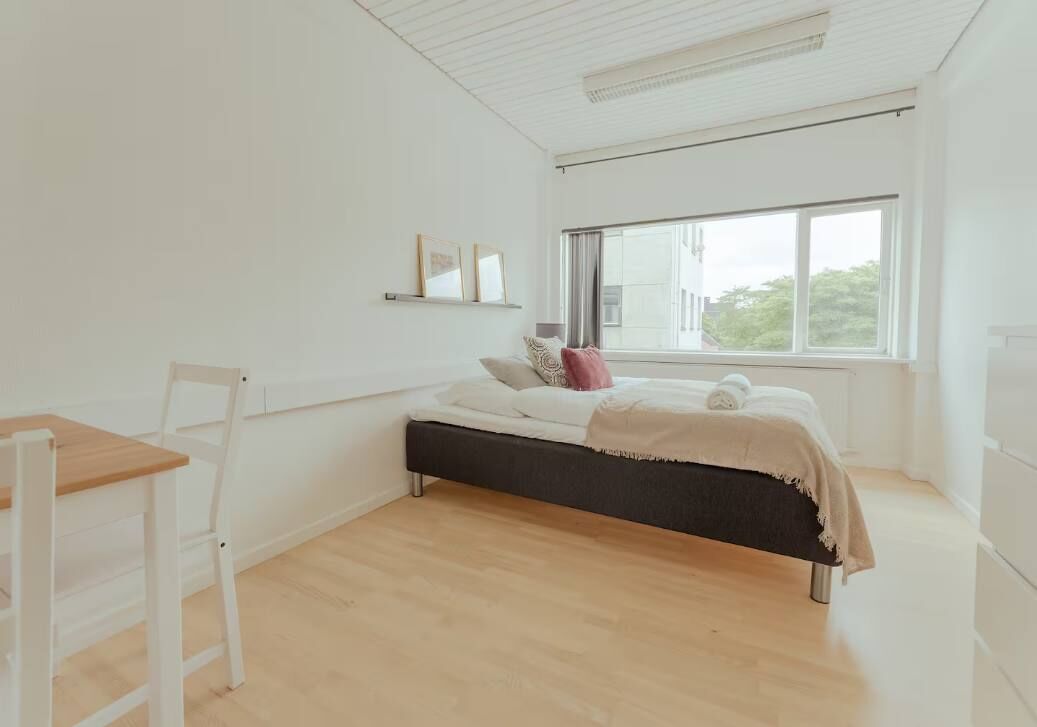 A bedroom with a bed, a chair, a window, and a shelf with pictures.
