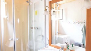 Un baño con ducha envuelta en vidrio, un lavamanos blanco con gabinete de madera y un espejo con luces empotradas.