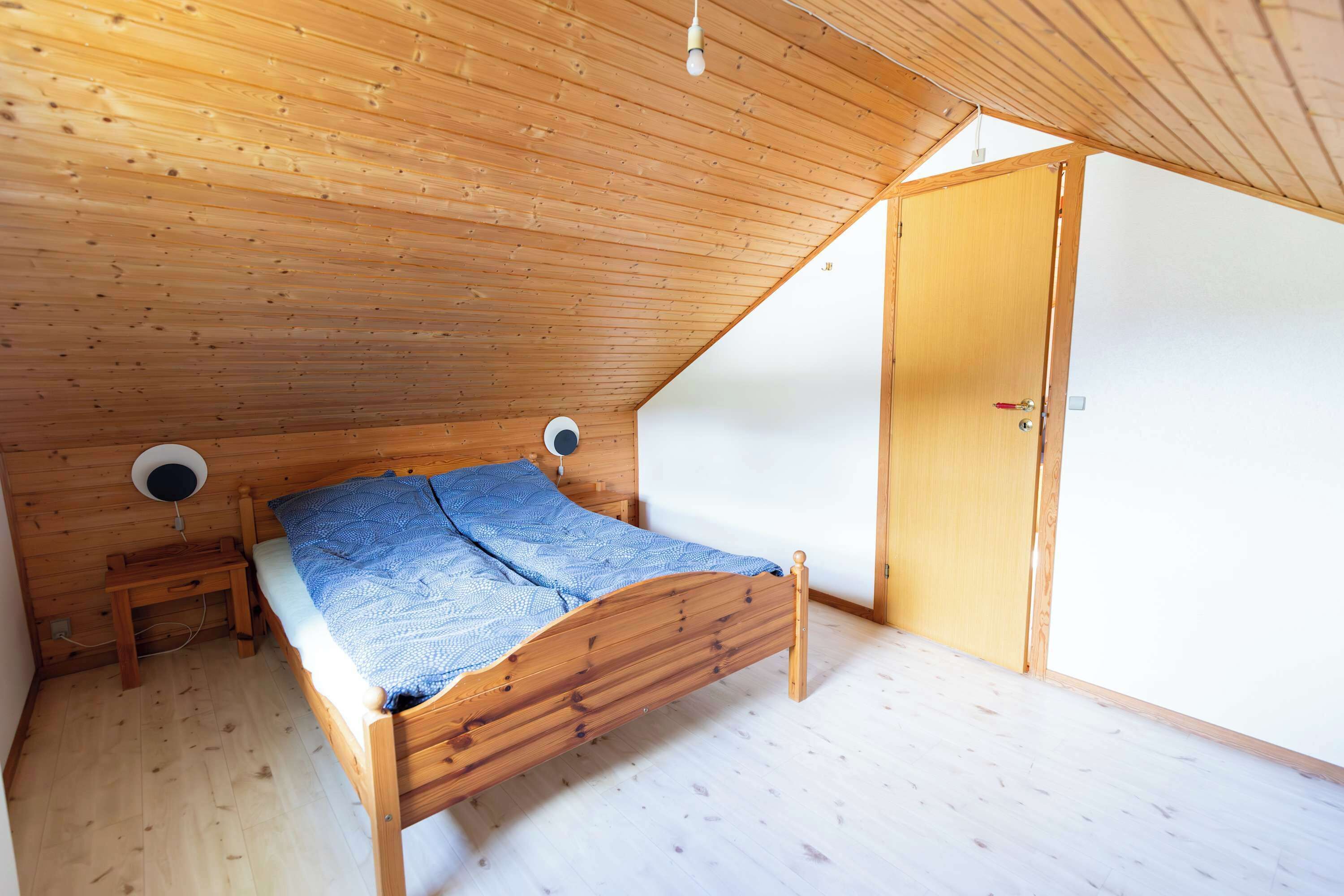 A single wooden bed in a room with a sloped ceiling, a small bedside table, and a door.