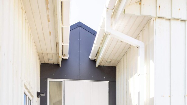 A narrow entrance with a wooden staircase leading to a white door, a trash bin to the left, and a metal step at the door's base.
