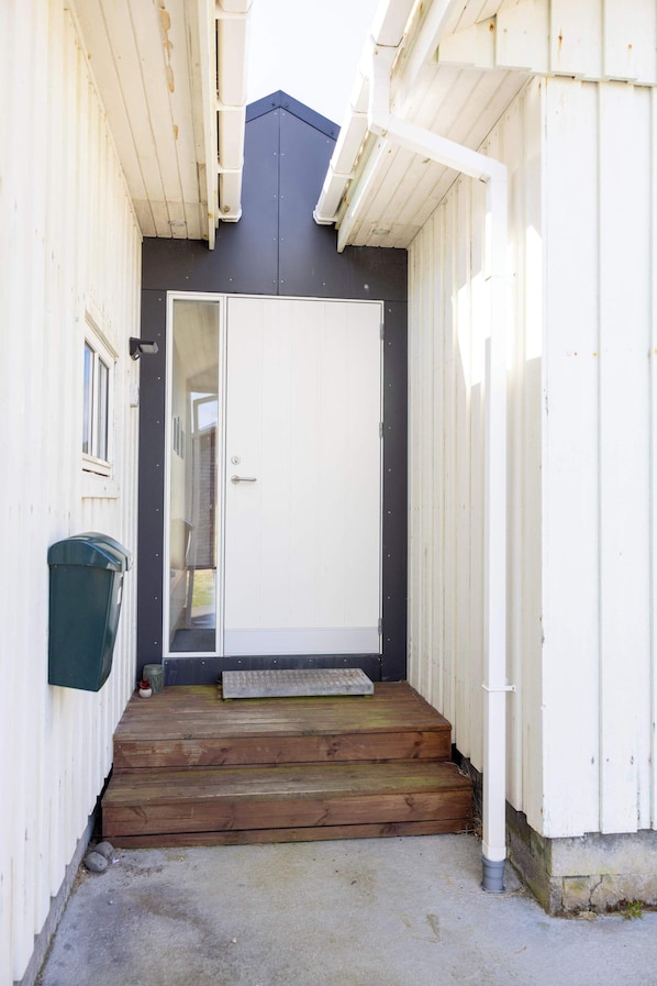 A narrow entrance with a wooden staircase leading to a white door, a trash bin to the left, and a metal step at the door's base.