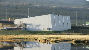 Un bâtiment moderne doté d’un toit plat et d’une grande surface réfléchissante, situé à proximité d’une étendue d’eau et d’une zone herbeuse.