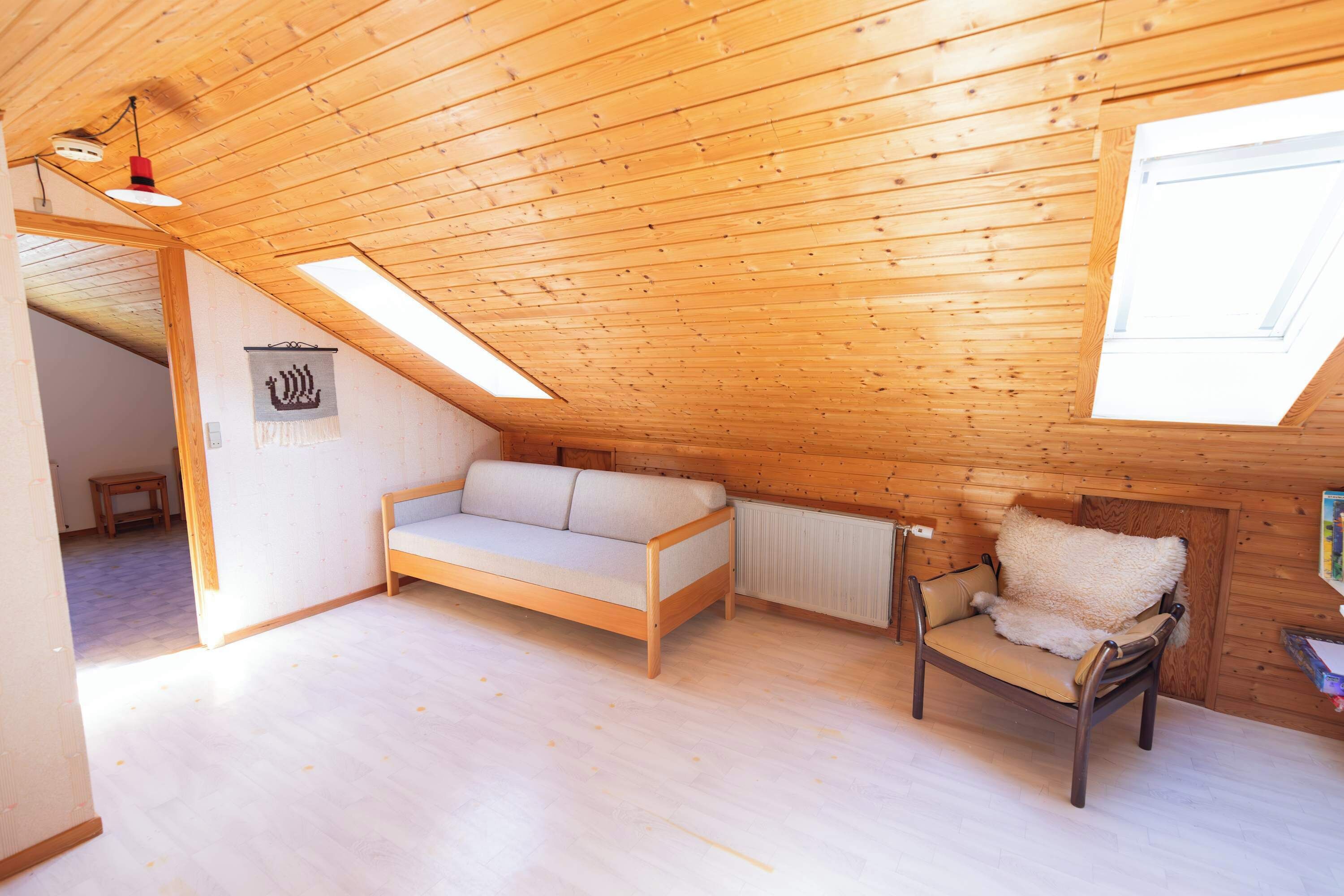 Ein gemütliches Dachzimmer mit Holzbalkendecke, einer Couch, einem Sessel und einer Heizung.