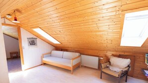 A cozy attic room with a wooden sloped ceiling, a sofa, a chair, and a radiator.