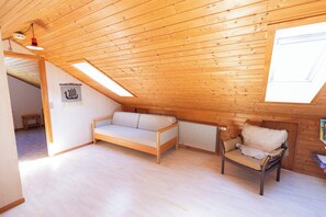 A cozy attic room with a wooden sloped ceiling, a sofa, a chair, and a radiator.