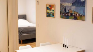 Een eetgedeelte met een houten tafel en witte stoelen, een schilderij aan de muur, en een bed op de achtergrond.