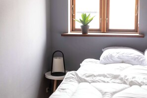 Un dormitori amb llit, una petita taula de nit, una planta en test i una finestra amb emmarcament de fusta.