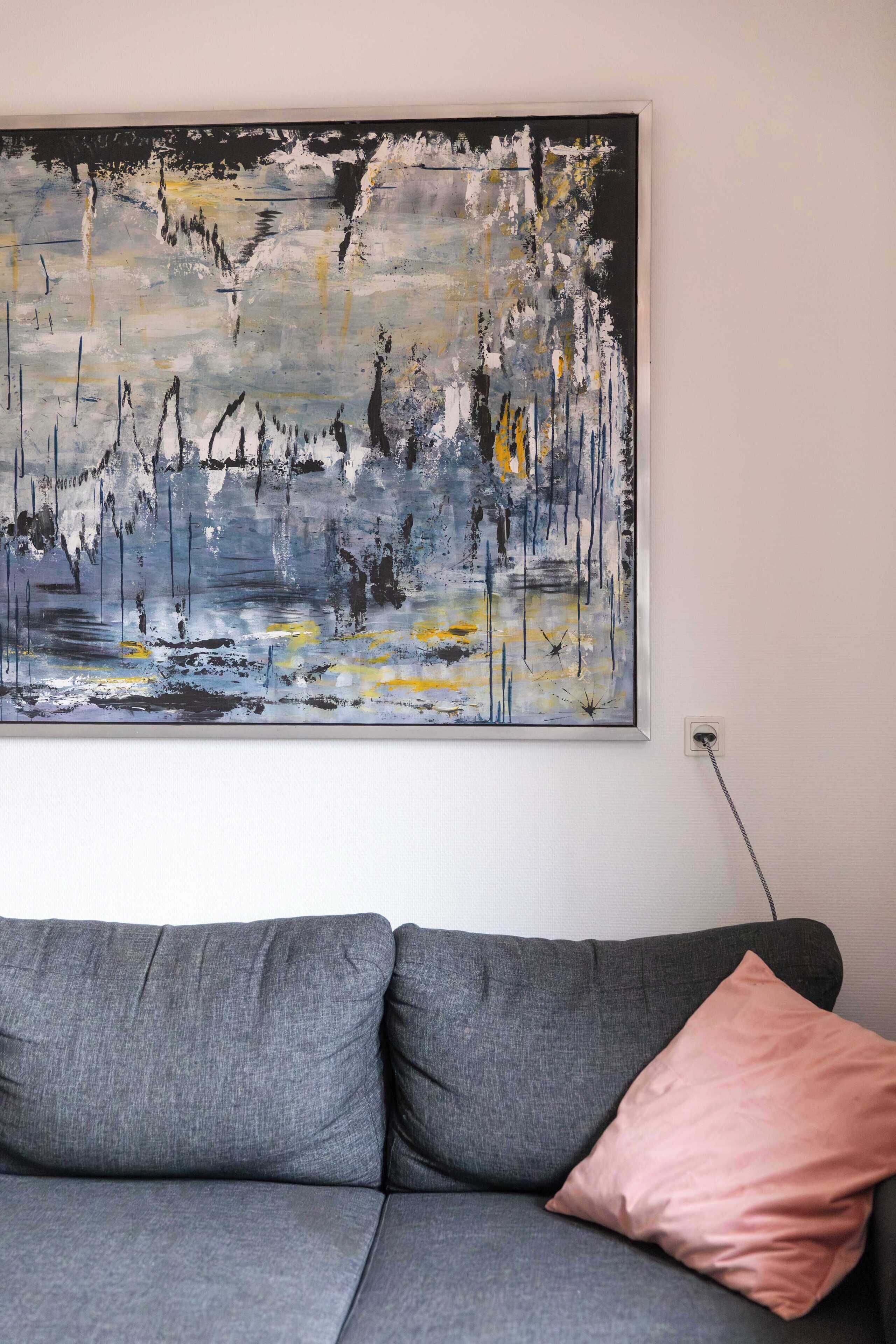 En grå sofa med en enkelt lyserød pude foran en hvid væg, med et stort abstrakt maleri over sofaen.