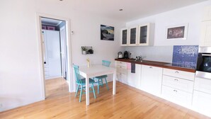Une cuisine compacte avec des meubles blancs, un plan de travail en bois et un coin repas comprenant une table blanche et des chaises turquoise.