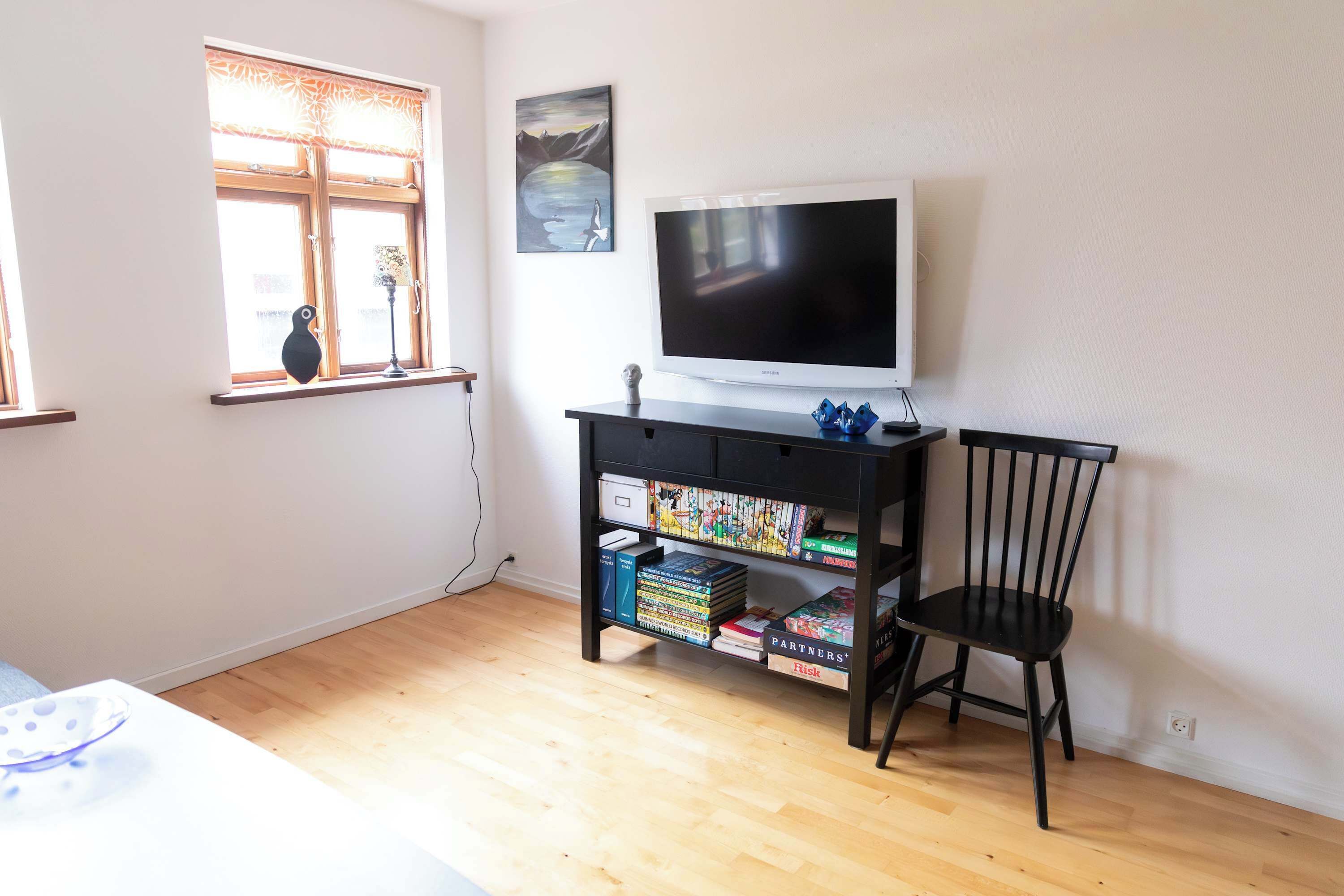 Ein Zimmer mit Fernseher auf einem Schrank, einem Bücherregal mit Büchern und Spielen, einem Sessel und einem Fenster mit Bergblick.