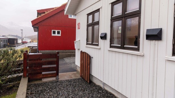 Un bâtiment blanc avec une grange rouge en arrière-plan, une porte en bois et une allée gravillonnée.