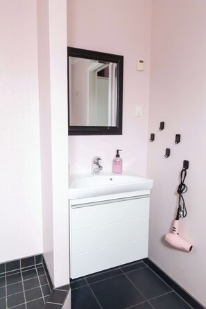 Een badkamer met een wit wastafelmeubel, een spiegel, een roze fles en een haardroger aan de muur.