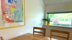 A dining area with a wooden table and chairs, a framed abstract painting, a window with a view of greenery, and a potted plant on the windowsill. - Harbourfront Boathouse Vestmanna Bird Cliffs (Vestmanna)