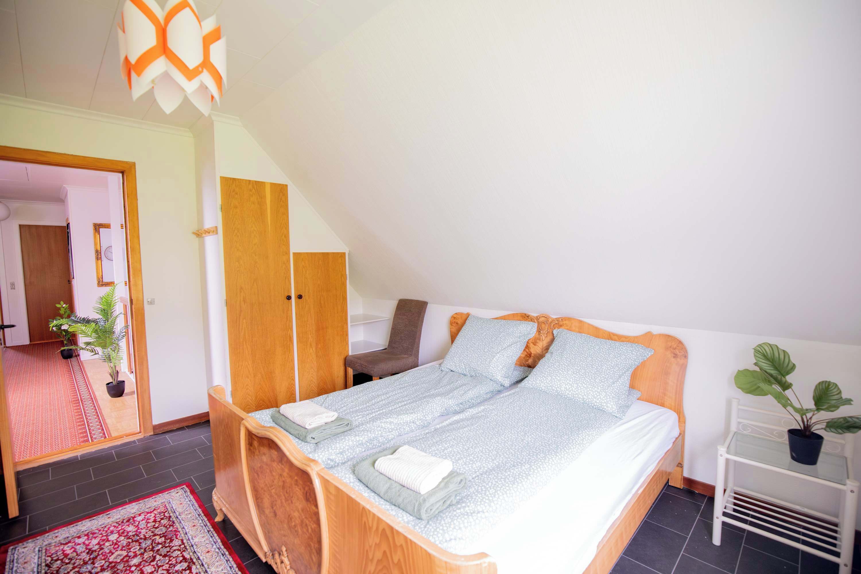 A bedroom with a bed, a chair, a wardrobe, a small table, and a potted plant.
