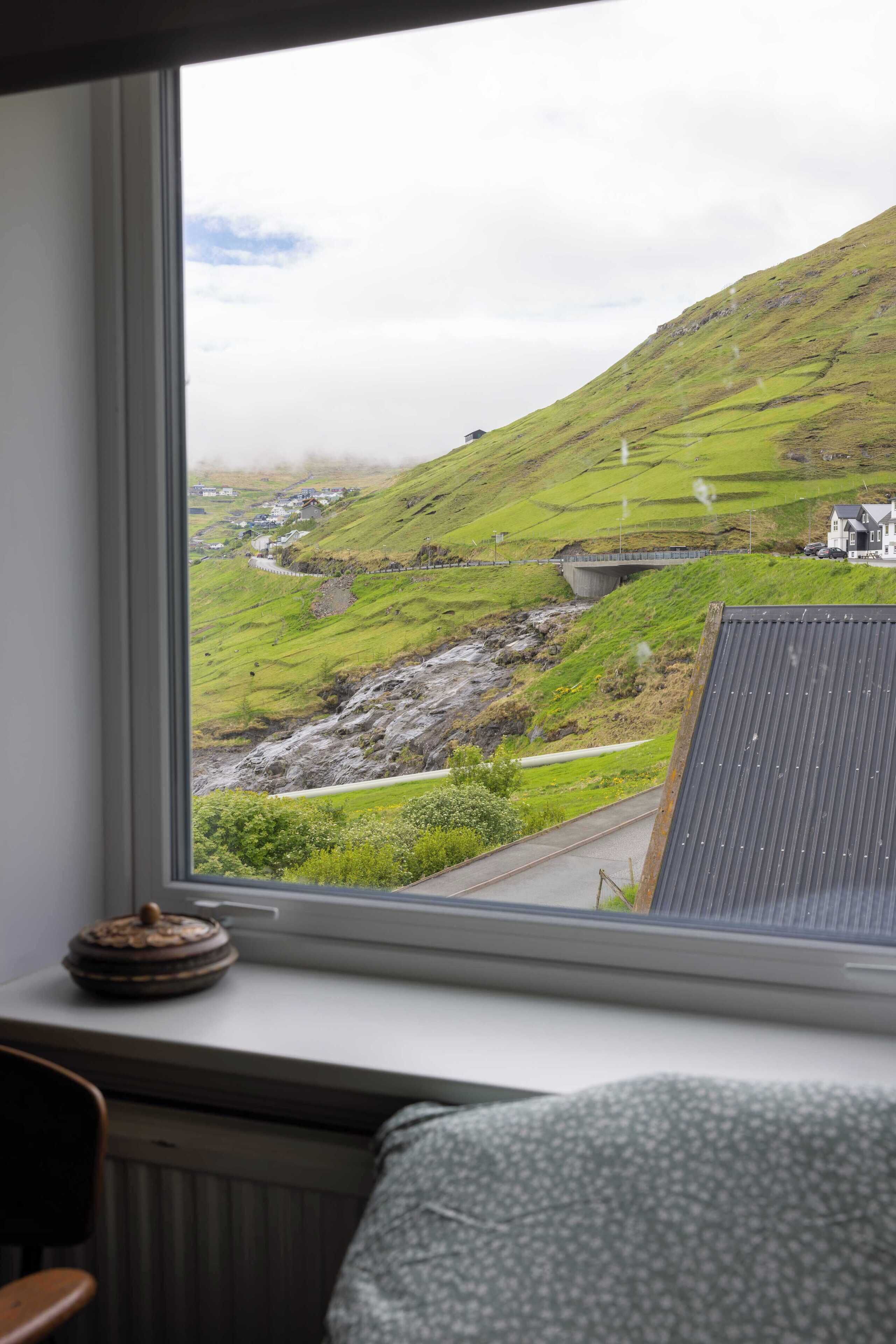 A window with a view of a green hillside, a bridge, and a small building.
