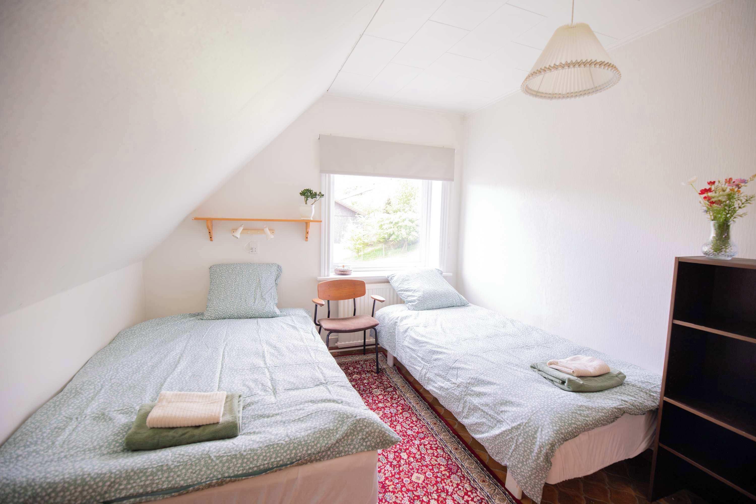 A small, well-lit bedroom with two single beds, a wooden chair, a small table, and a window providing natural light.