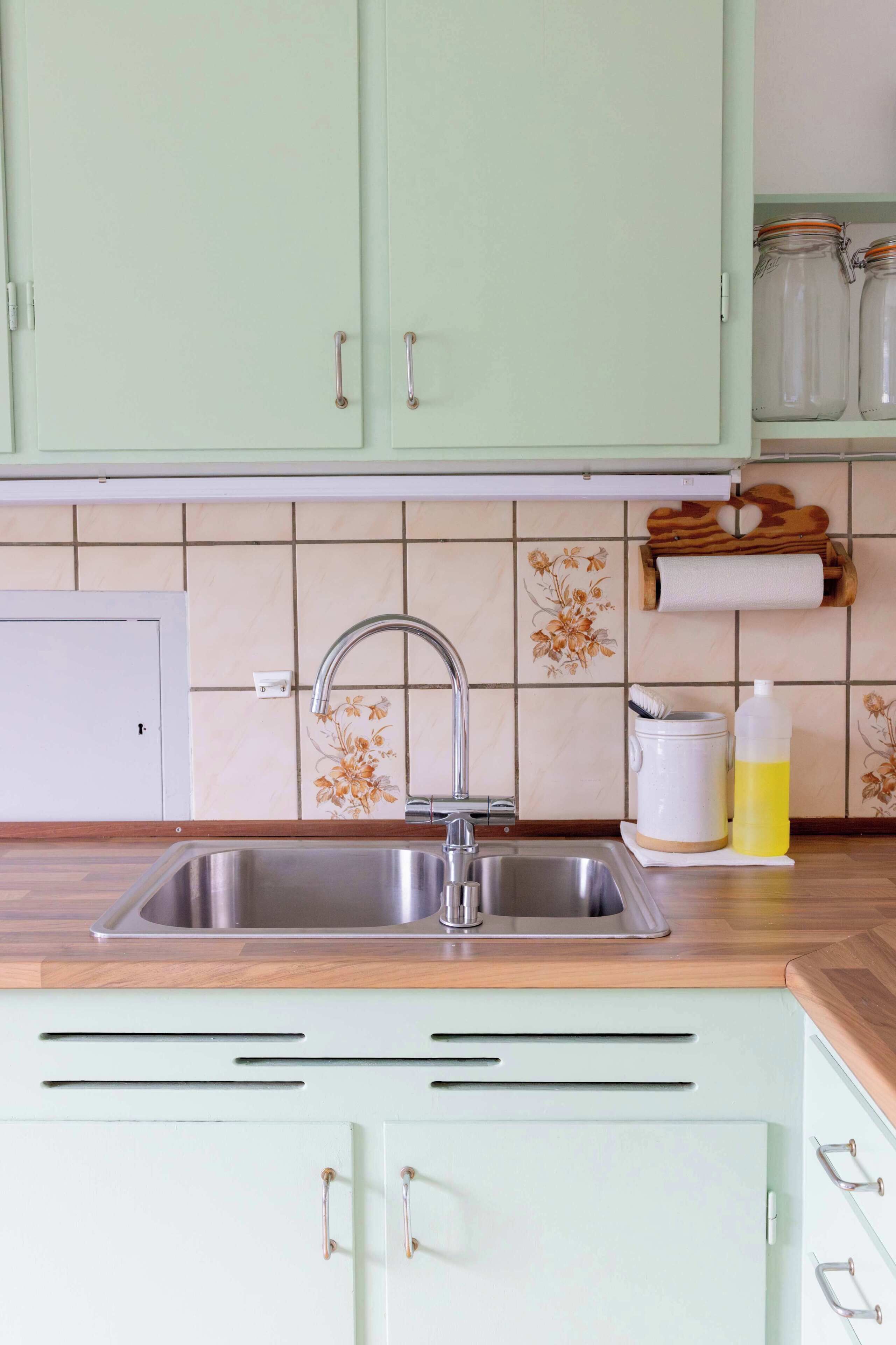 A kitchen with a stainless steel sink, light green cabinets, and a tiled backsplash with floral patterns.