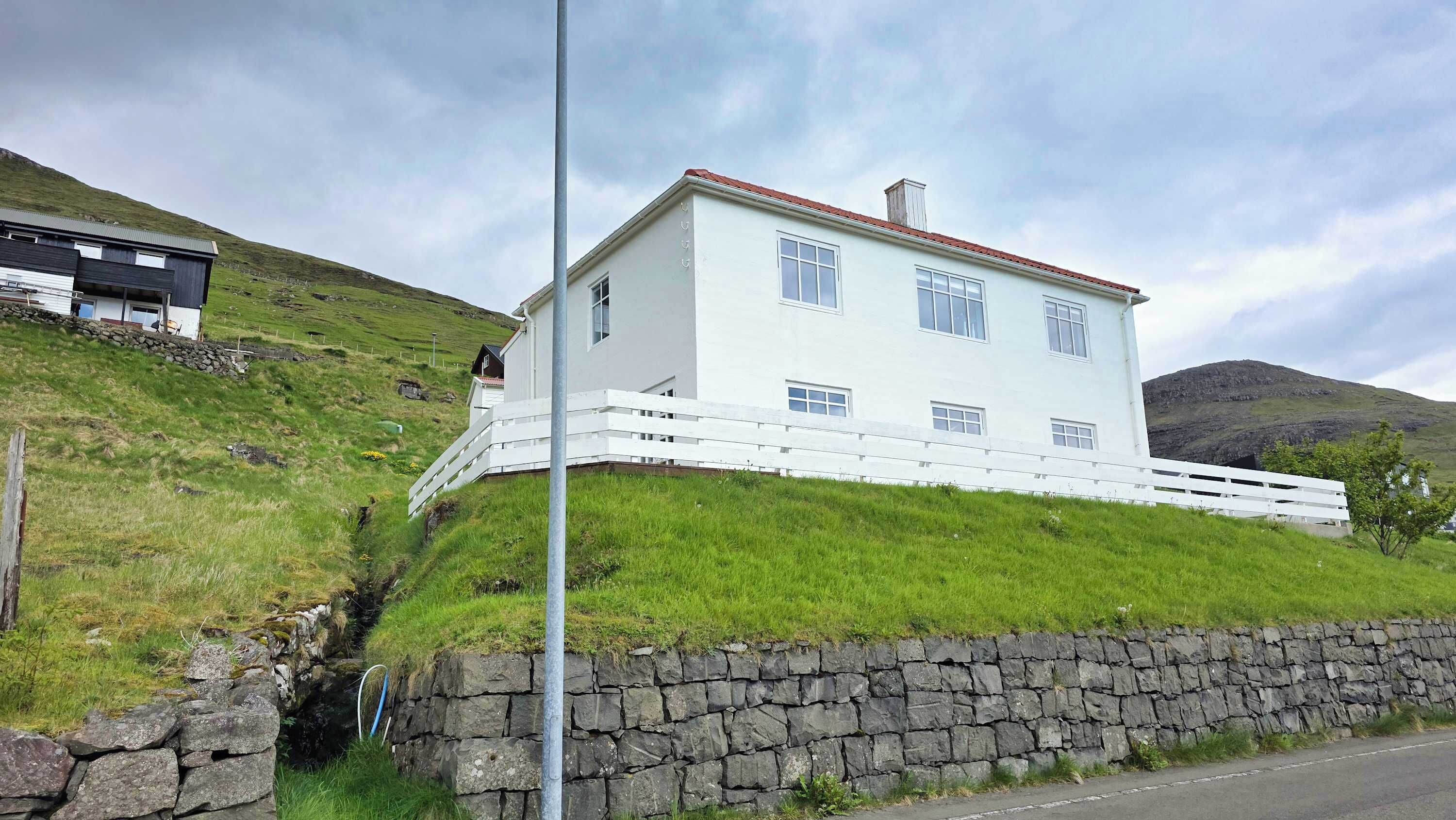 A white two-story house with a stone wall foundation, a white fence, and a grassy hillside.