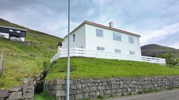 A white two-story house with a stone wall foundation, a white fence, and a grassy hillside.