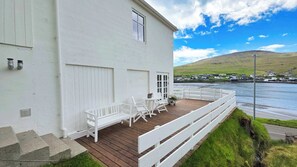 Un edificio blanco con terraza de madera, mobiliario exterior y vista a un cuerpo de agua y casas a lo lejos.