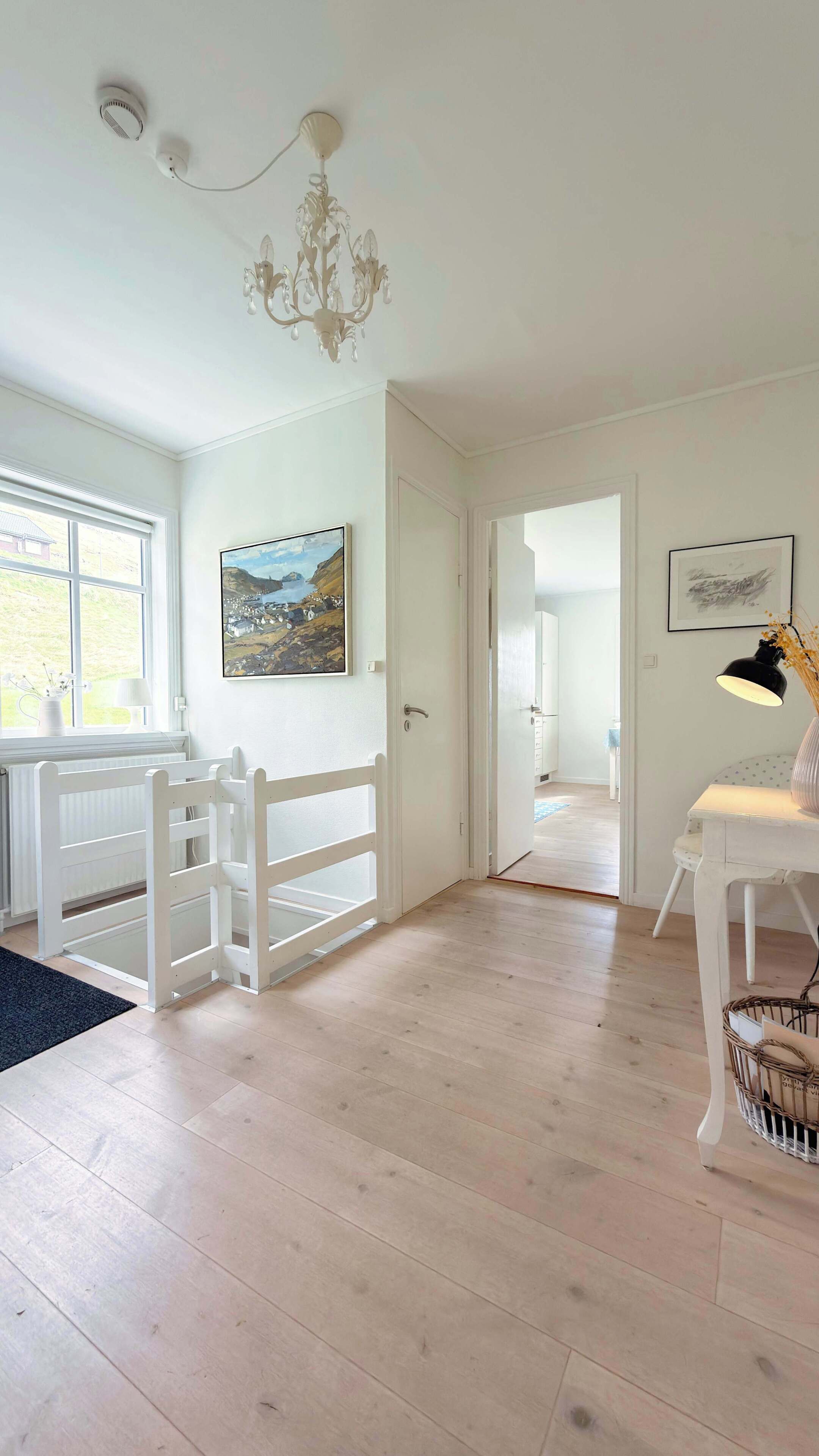 Una habitación con una puerta blanca, un candelabro, un cuadro, un escritorio con una lámpara y una cesta de mimbre.