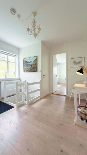 Una habitació amb una porta blanca, un candelabrum, un quadre, un escriptori amb una làmpada i una cistella de vimet.