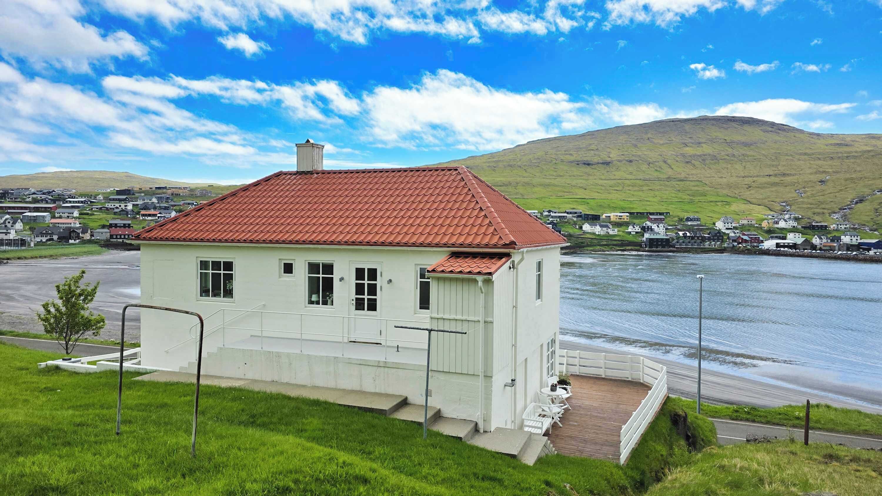 A white building with a red roof, situated near a body of water, with a view of a hilly landscape in the background.