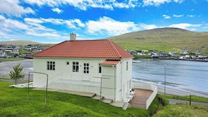 A white building with a red roof, situated near a body of water, with a view of a hilly landscape in the background.