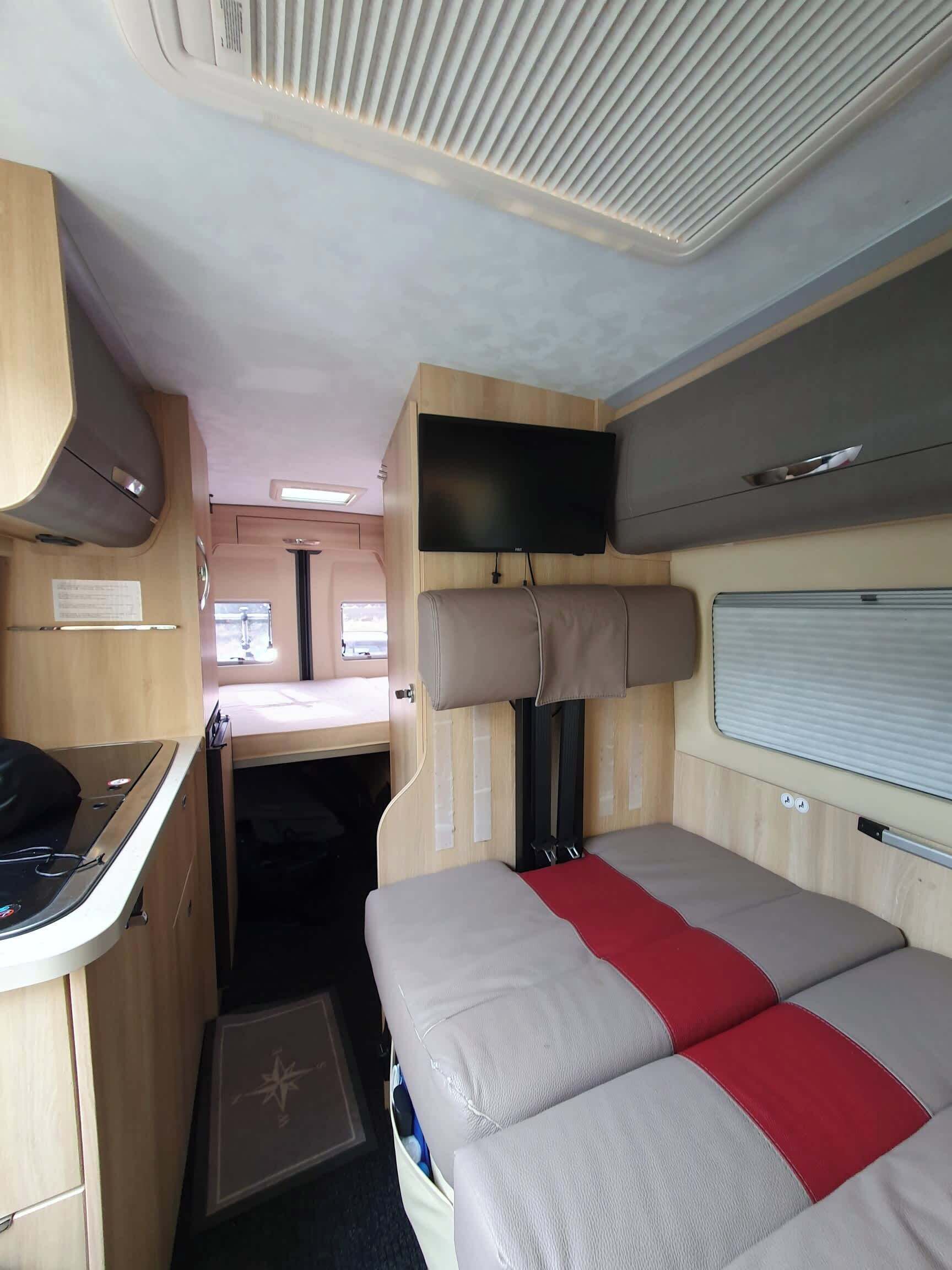A compact RV interior with a kitchenette, a bed with a red accent, a mounted TV, and a small window.