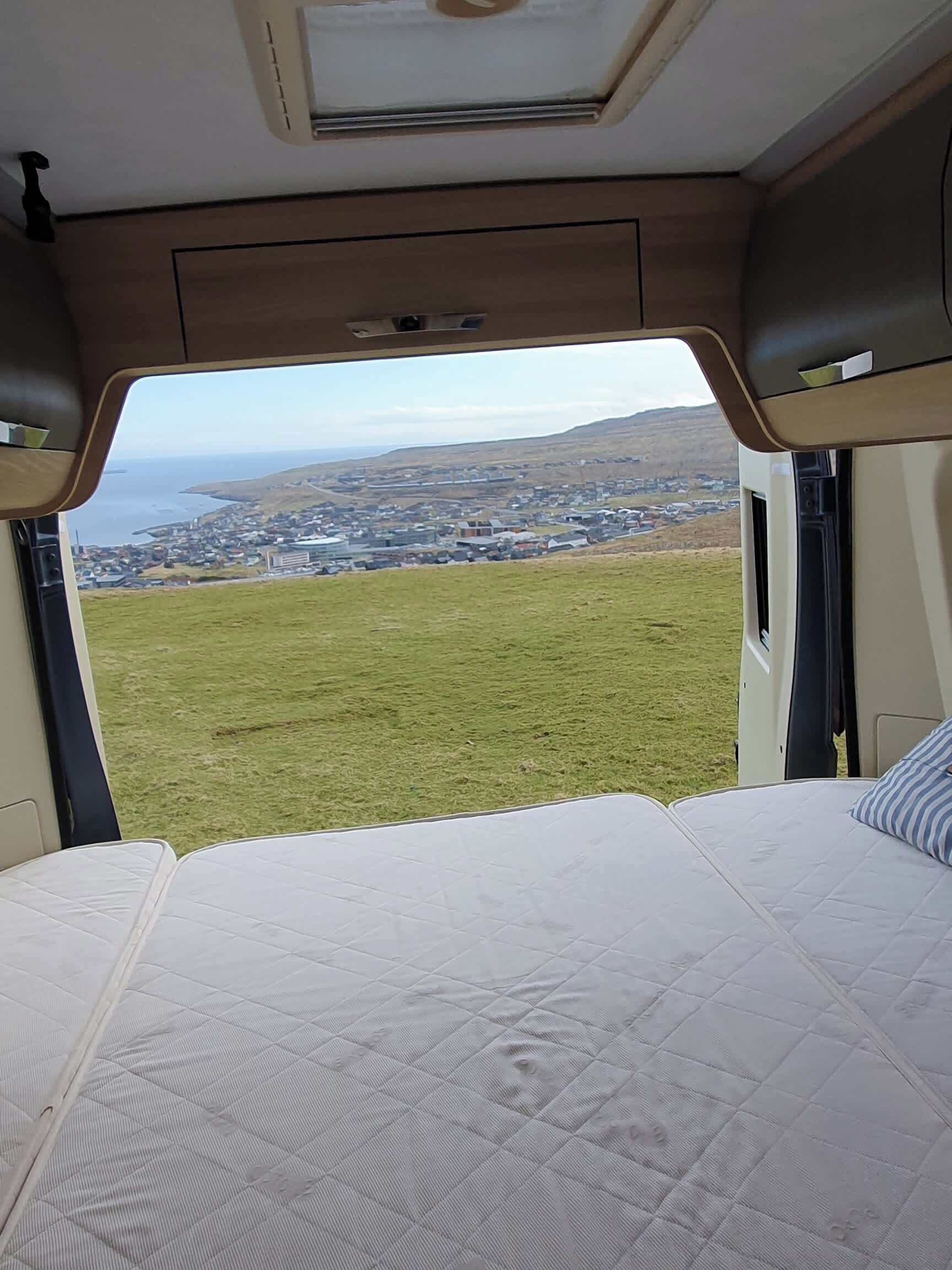 Interno di un camper con un letto e vista su un prato e degli edifici.