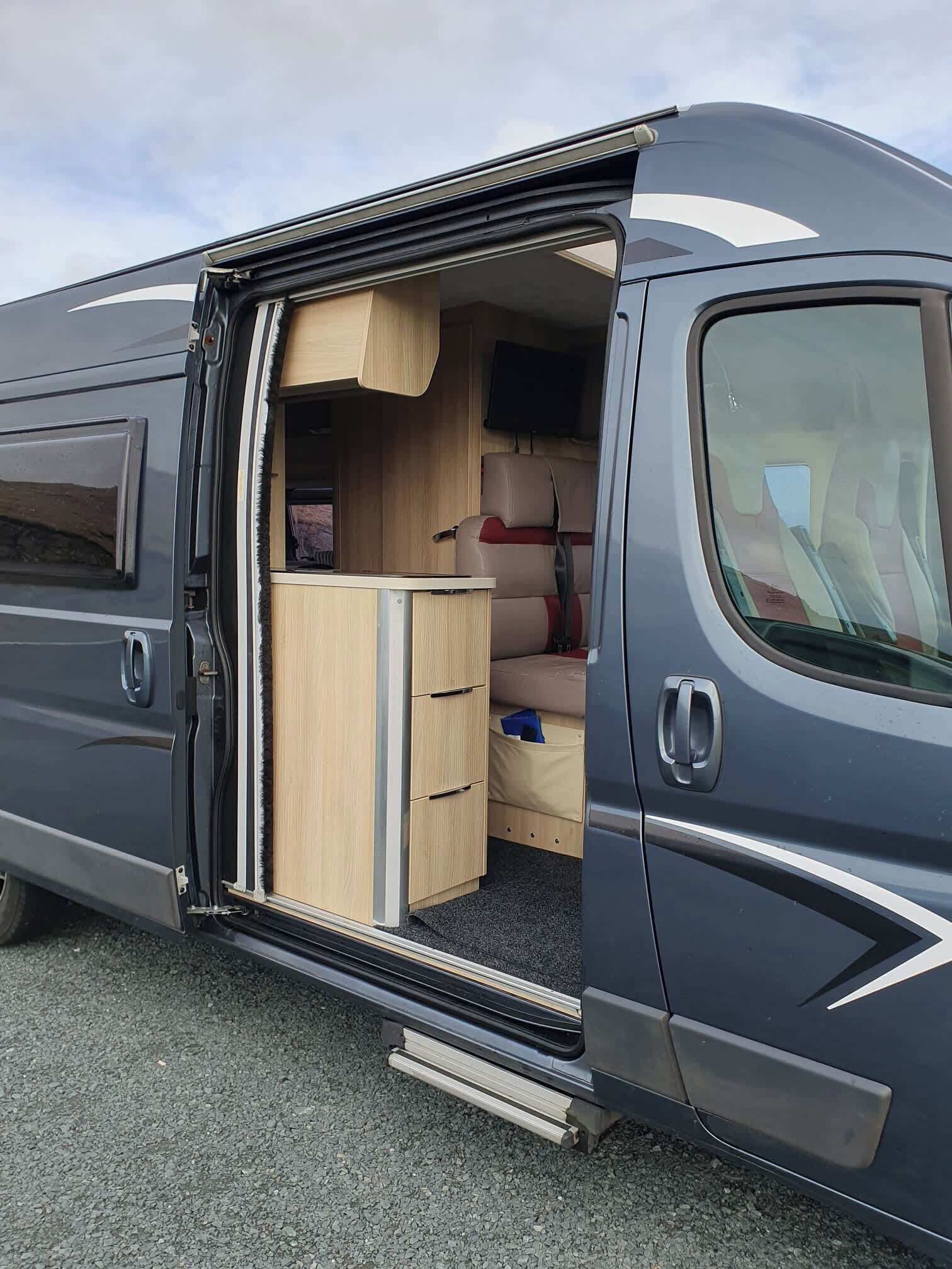 A van with a sliding door open, revealing a compact kitchenette with a sink, a small refrigerator, and a wooden cabinet.