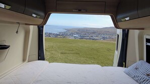 A campervan interior with a bed, a view of a town and hills, and a window. - Adventure Autocamper Van to Explore the Faroes (Hoyvík)