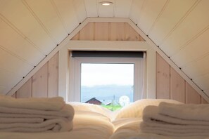 Une pièce avec un plafond en pente, une fenêtre donnant sur un paysage et un lit recouvert de draps blancs pliés.
