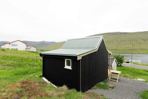 Une petite construction en bois d’un seul étage, dotée d’un toit en pente, située sur une colline herbeuse, avec un plan d’eau et des collines en arrière-plan.