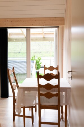 A dining area with a table and chairs, a view of a green landscape through a large window, and a wooden ceiling.