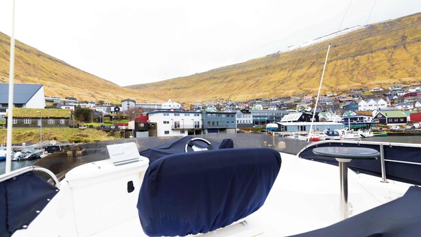 A boat's deck with a covered seating area, a steering wheel, and a Garmin device. In the background, there is a small town with houses and boats docked along the waterfront.