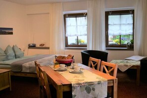 Ein gemütliches Zimmer mit einem Bett, einem Esstisch mit Stühlen und zwei Fenstern mit Vorhängen.