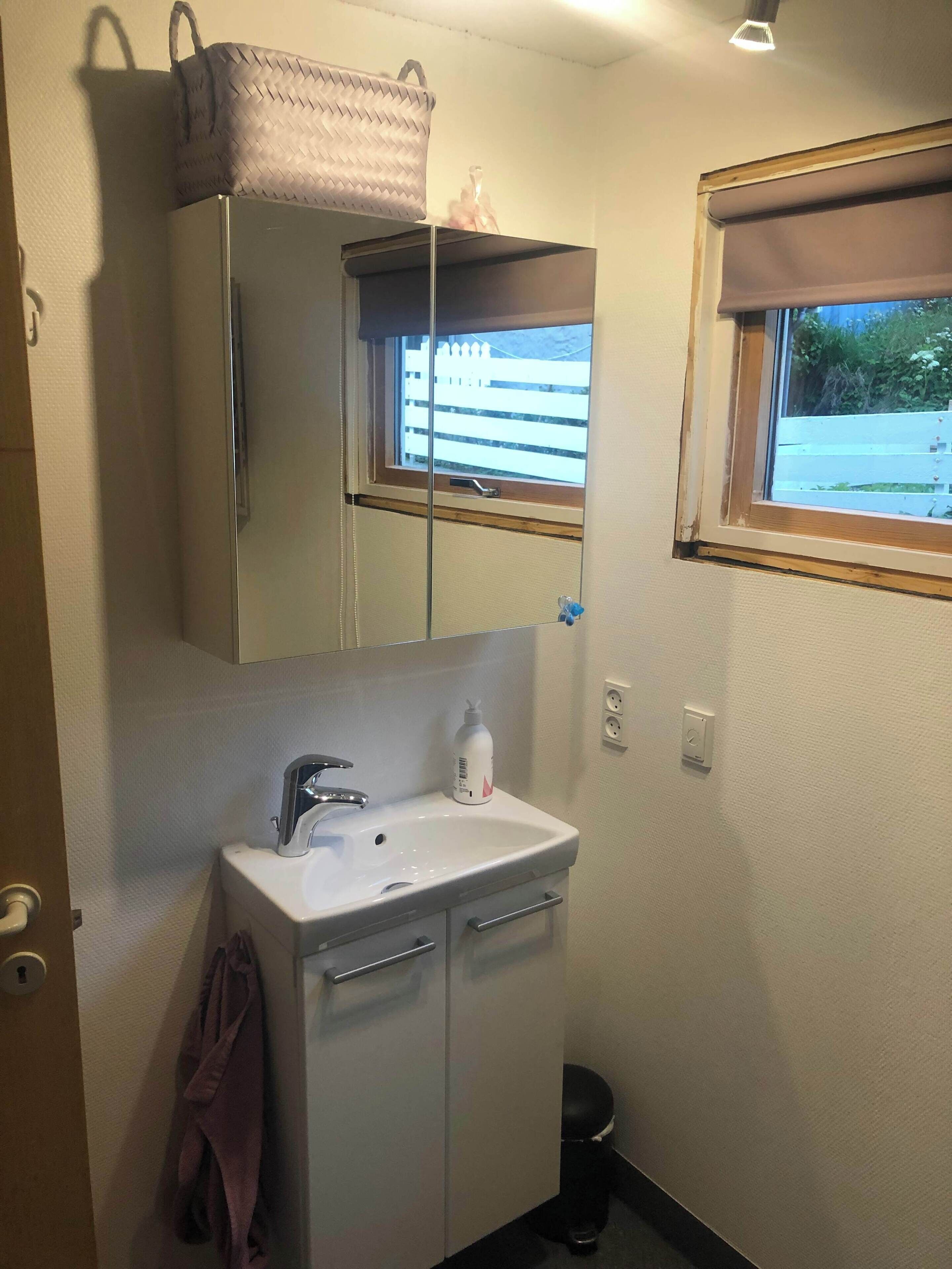 Une petite salle de bain avec un lavabo blanc, un miroir, une fenêtre donnant sur une clôture et une serviette accrochée au mur.