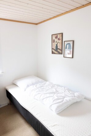 A single bed with white bedding in a room with white walls and a wooden ceiling. There are two framed pictures on the wall.