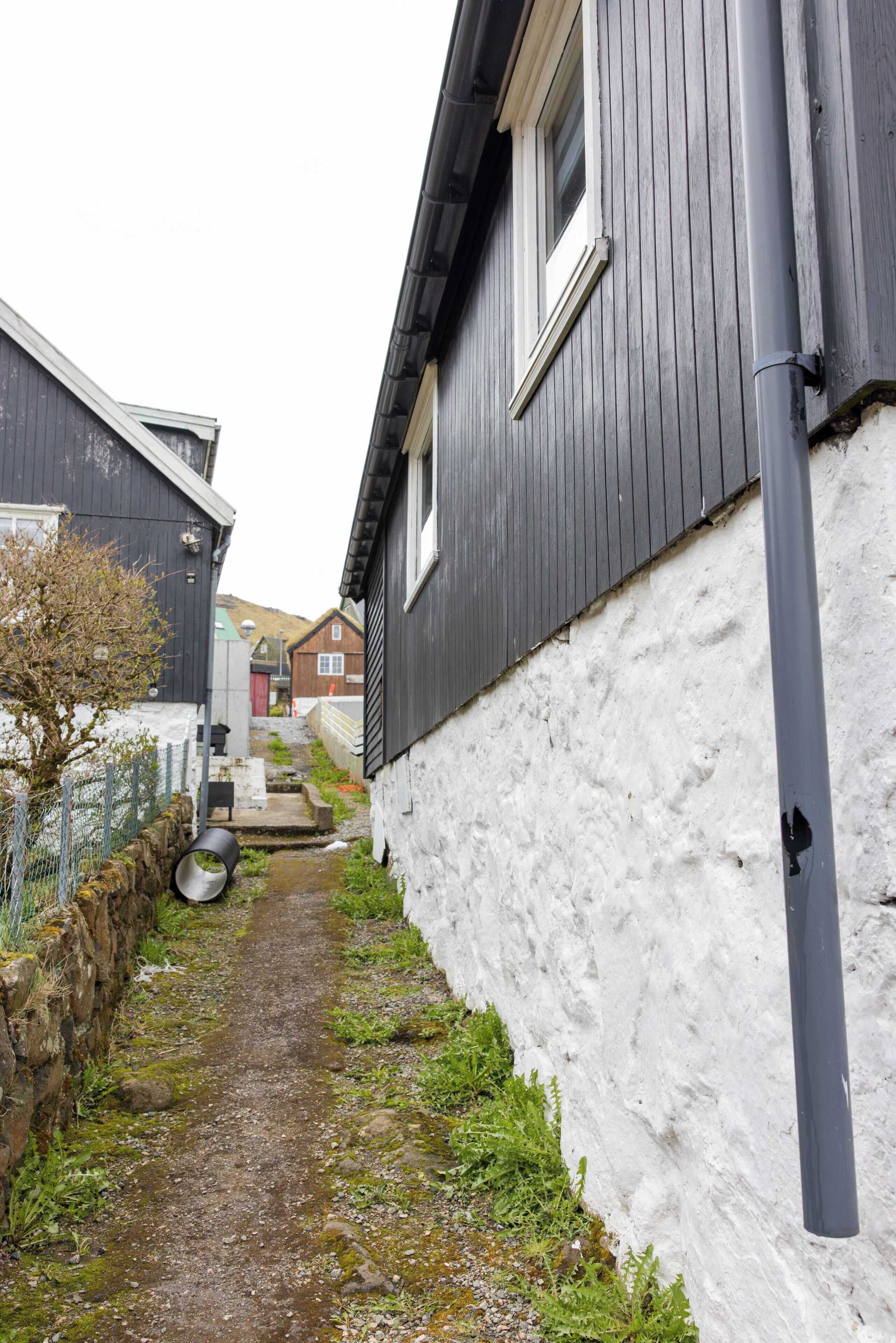 Ein schmaler Weg zwischen Gebäuden, mit einer weißen Wand auf der einen Seite und einem dunklen Holzhaus auf der anderen.
