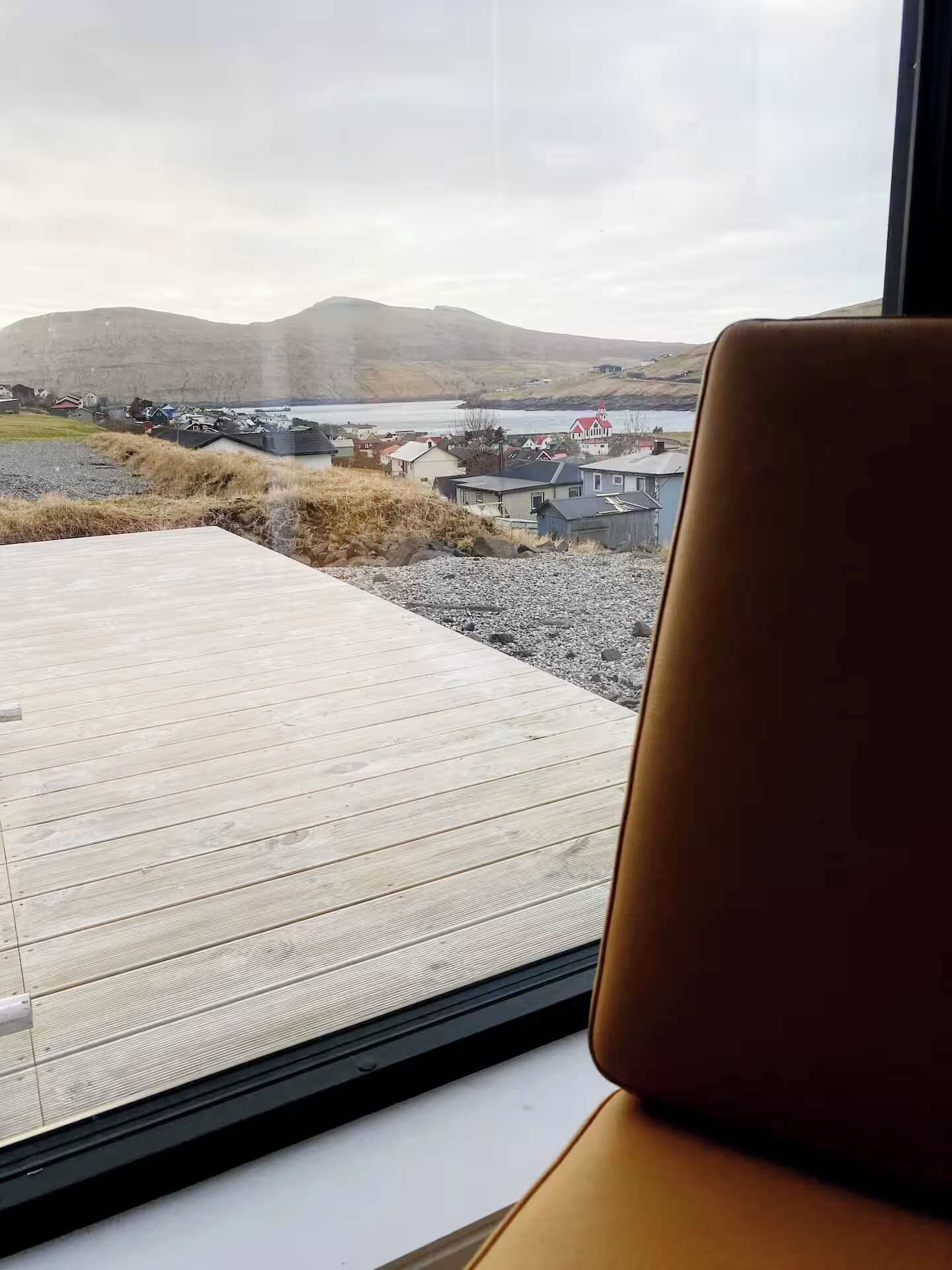 A view from a window with a wooden deck and a chair, overlooking a rural landscape with houses and a body of water.