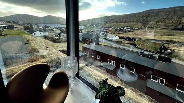 Une pièce dotée d’une grande fenêtre donnant sur un paysage rural, avec des maisons, un plan d’eau et des collines.